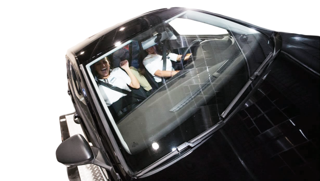 Widok z przodu, przez szybę samochodu, ukazuje dwie osoby siedzące na przednich fotelach. Ubrane w białe koszule, zapięte w pasy, uczestniczą w symulacji jazdy lub demonstracji bezpieczeństwa. Kierowca skupiony, kontroluje pojazd; pasażer reaguje z zaskoczeniem lub ekscytacją — to moment, który pokazuje, jak intensywne może być doświadczenie edukacyjne.