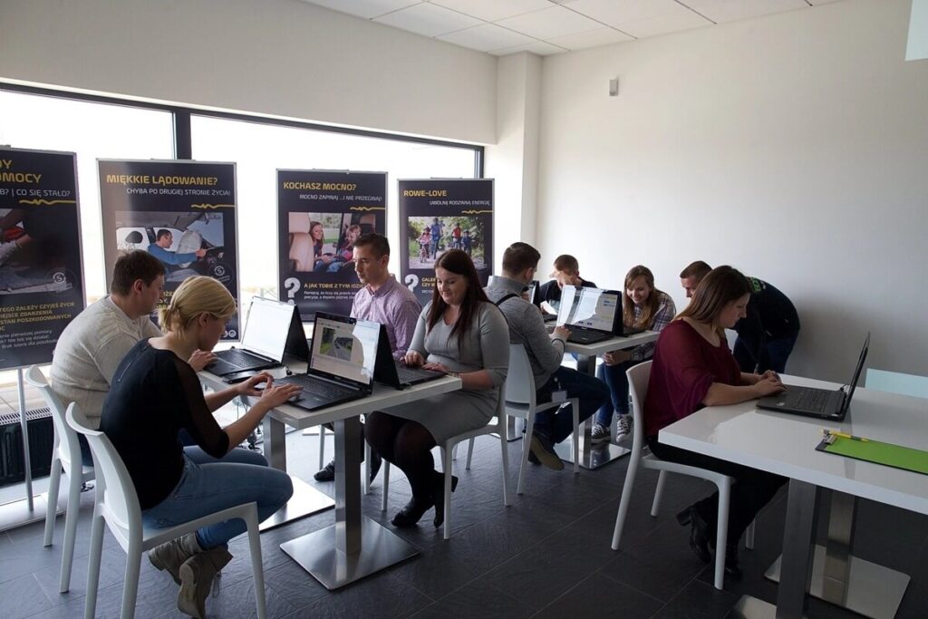 Na zdjęciu widać grupę uczestników biorących udział w warsztatach szkoleniowych w nowoczesnej sali. Kilka osób siedzi przy stolikach i pracuje na laptopach, wykonując zadania lub analizując materiały edukacyjne. Uczestnicy są skupieni i zaangażowani w pracę, co sugeruje praktyczny charakter szkolenia.