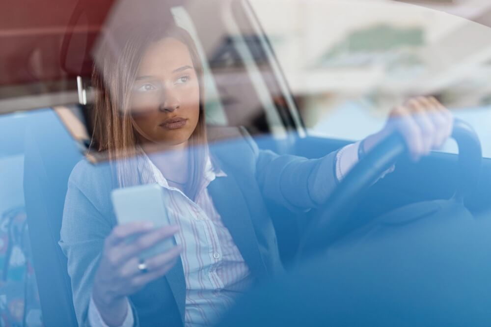 Na zdjęciu widać kobietę prowadzącą samochód, która jednocześnie trzyma w dłoni telefon. Ujęcie wykonano z zewnątrz pojazdu, przez szybę, co podkreśla temat korzystania z telefonu podczas jazdy i zagrożeń związanych z rozproszeniem uwagi kierowcy.