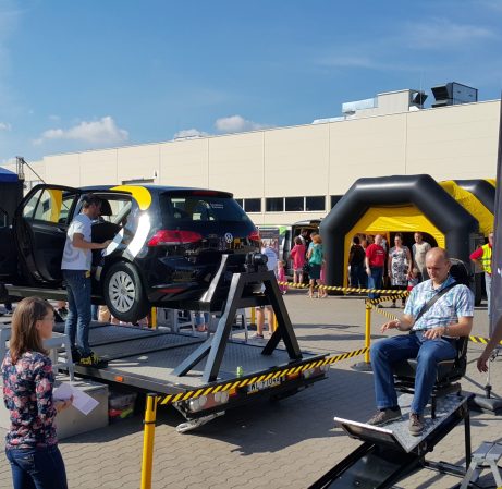 Na zdjęciu widzimy pokaz bezpieczeństwa drogowego zorganizowany w ramach wydarzenia „Świadomy Kierowca”. Główną atrakcją jest samochód zamontowany na platformie obrotowej, symulującej dachowanie, co ma na celu pokazanie, jak działają pasy bezpieczeństwa podczas wypadku. W tle znajdują się żółte dmuchane konstrukcje, obserwatorzy oraz urządzenie imitujące siłę uderzenia, wszystko w otoczeniu taśmy ostrzegawczej, podkreślającej edukacyjny i kontrolowany charakter demonstracji.